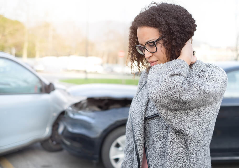 Standing next to two cars that have crashed into each other, a woman puts her hand on the back of her neck. Car accidents are among the personal injury cases that the Alabama injury attorneys at The Clarkson Firm takes.