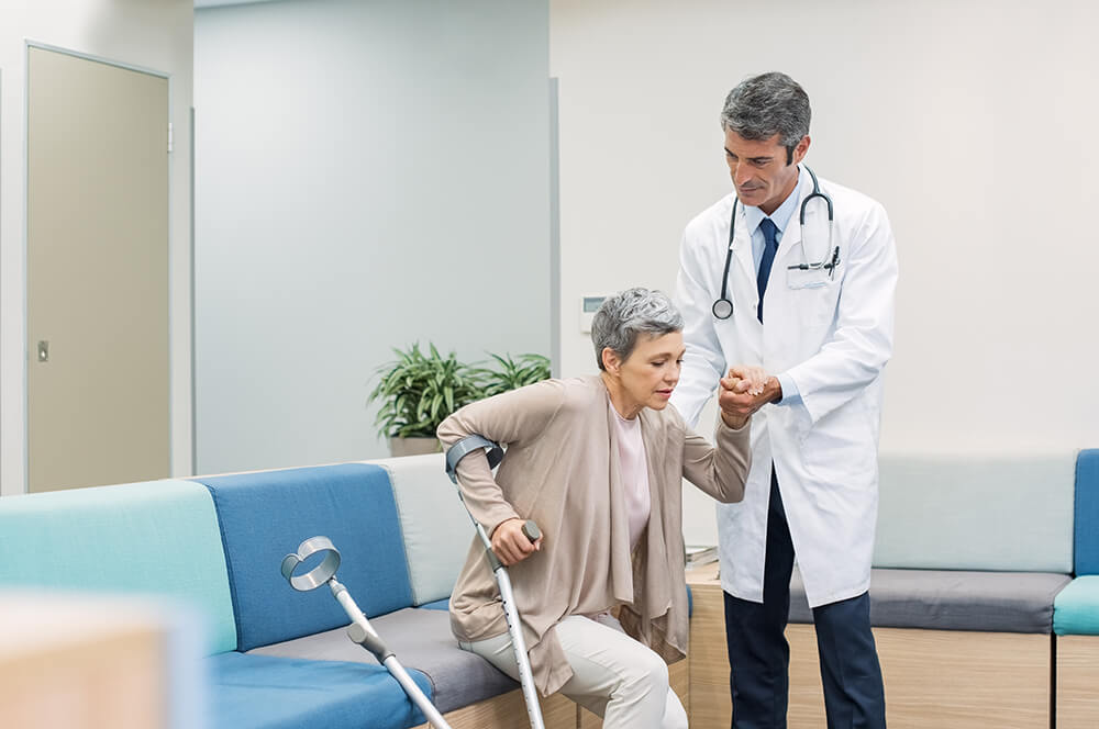 A doctor helps a woman with crutches stand up from a waiting room seat. Many medical impairments can meet the qualifications for Social Security Disability benefits.