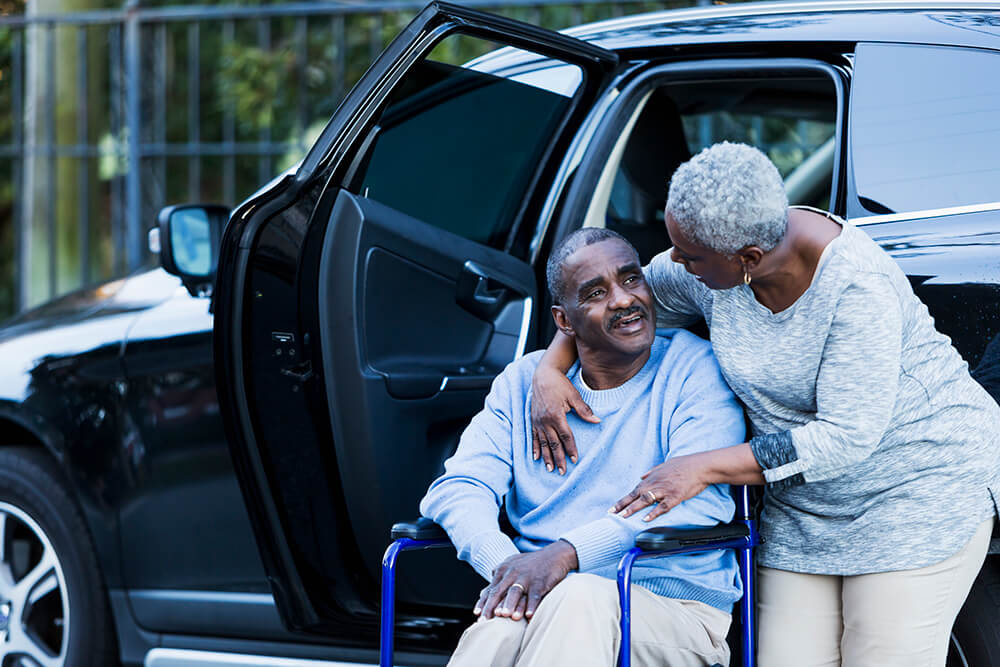 A woman stands behind a man in a wheelchair, hugging him after they get out of a car. SSI eligibility includes financial and health requirements.
