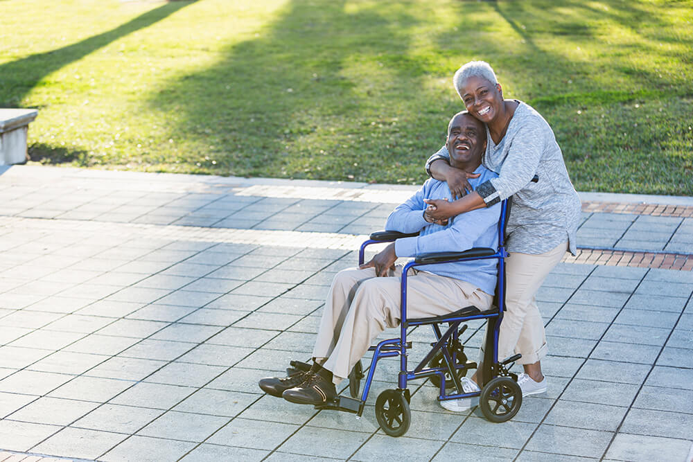 Sitting in a wheelchair in a park, a man smiles as a woman standing behind him give him a hug. A disability lawyer can help with the SSI appeals process.