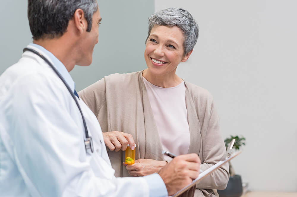Holding a prescription bottle, a woman smiles and talks to a doctor with a clipboard. Information about your medical treatment is part of what you need to qualify for Social Security Disability.