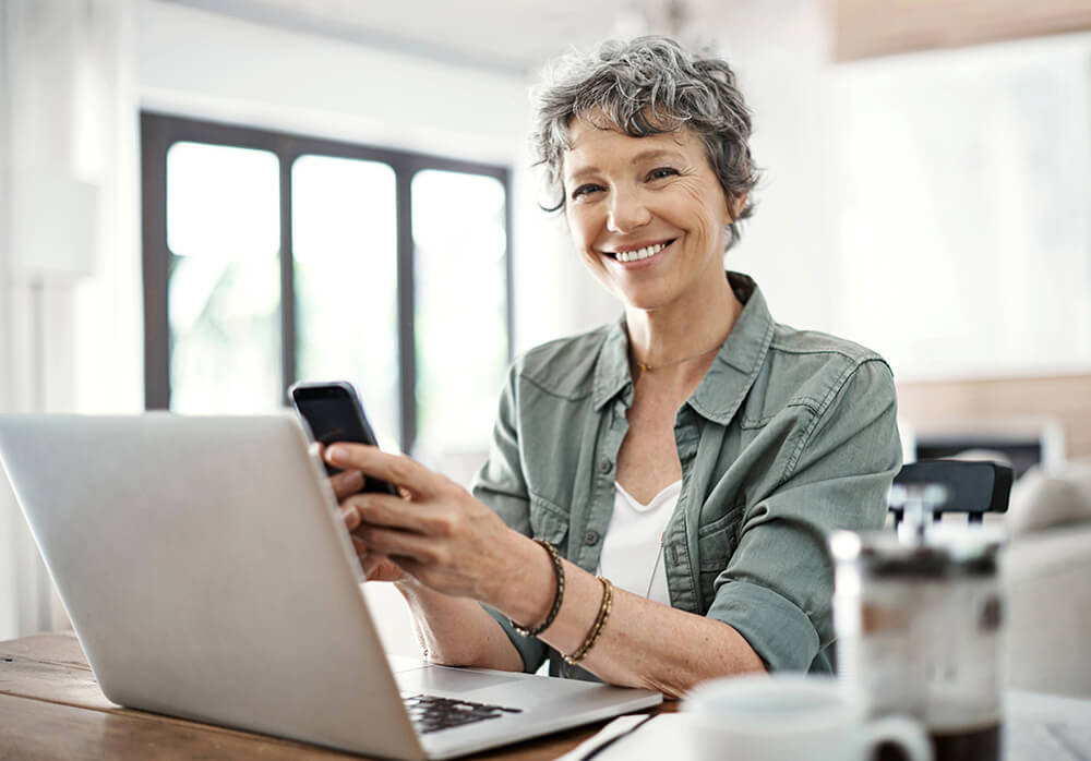 Laptop open in front of her, a woman holds her smartphone and smiles. The Clarkson Firm disability attorneys can answer questions about getting Social Security Disability benefits.