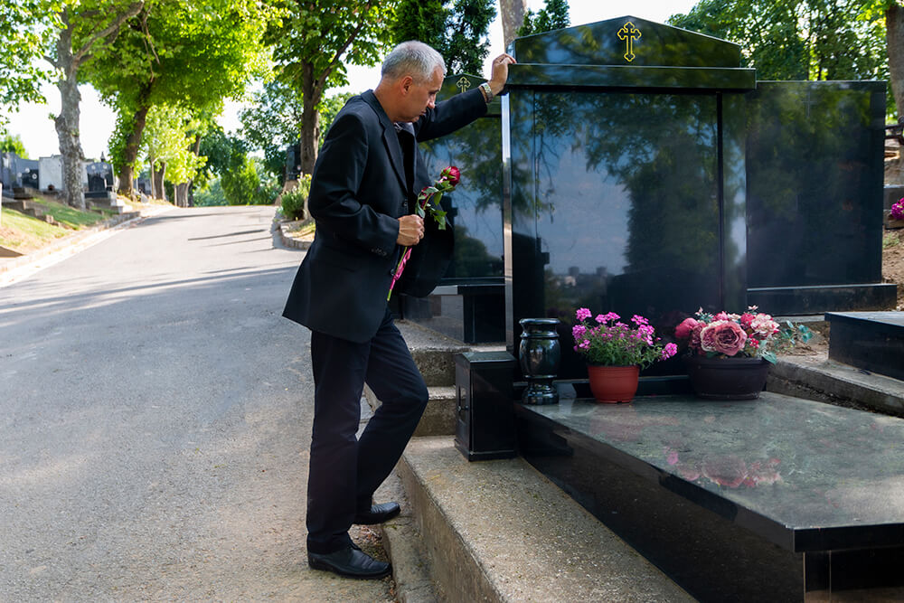 Wearing a suit and holding a flower, a man puts one hand on a gravestone and looks down. In a funeral home negligence case, you can claim various forms of compensation.