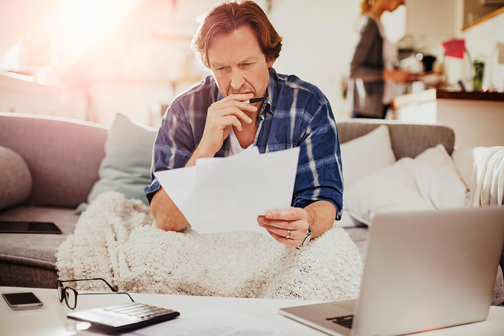 With a blanket over his legs, a man sits on a couch studying paperwork. You can be denied SSD benefits for multiple reasons.