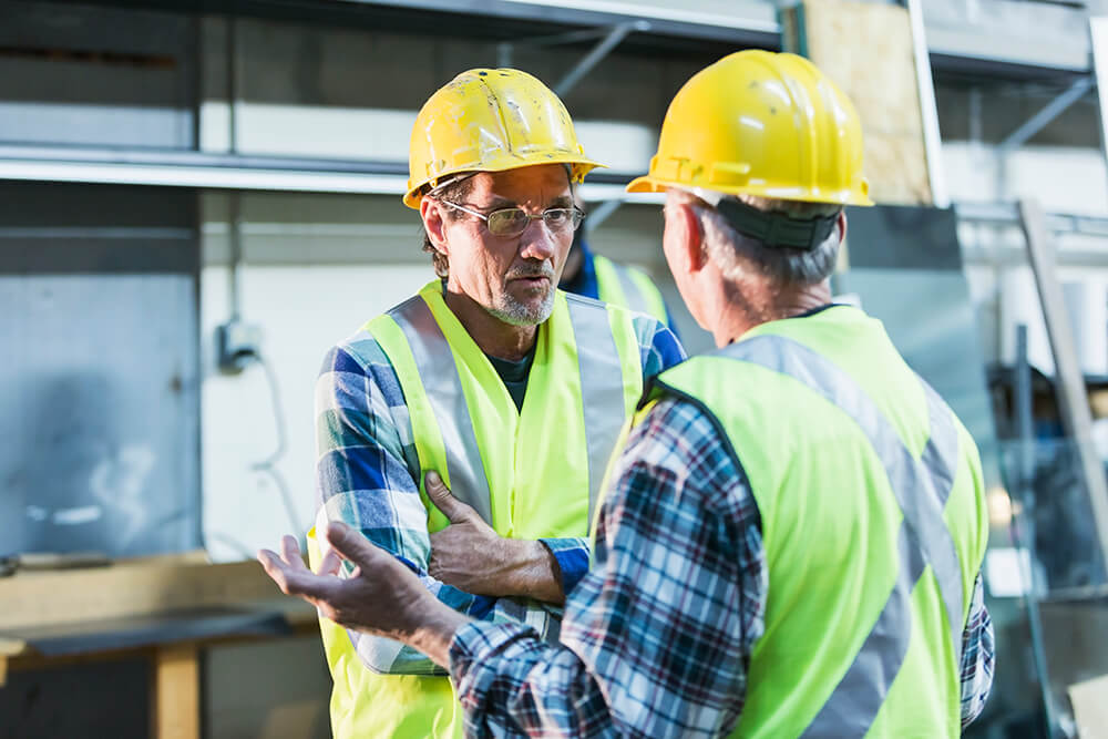In a workplace setting, two men wearing safety vests and hardhats talk to each other. The Alabama disability lawyers at The Clarkson Firm can point people to resources for getting by while waiting for disability benefits.