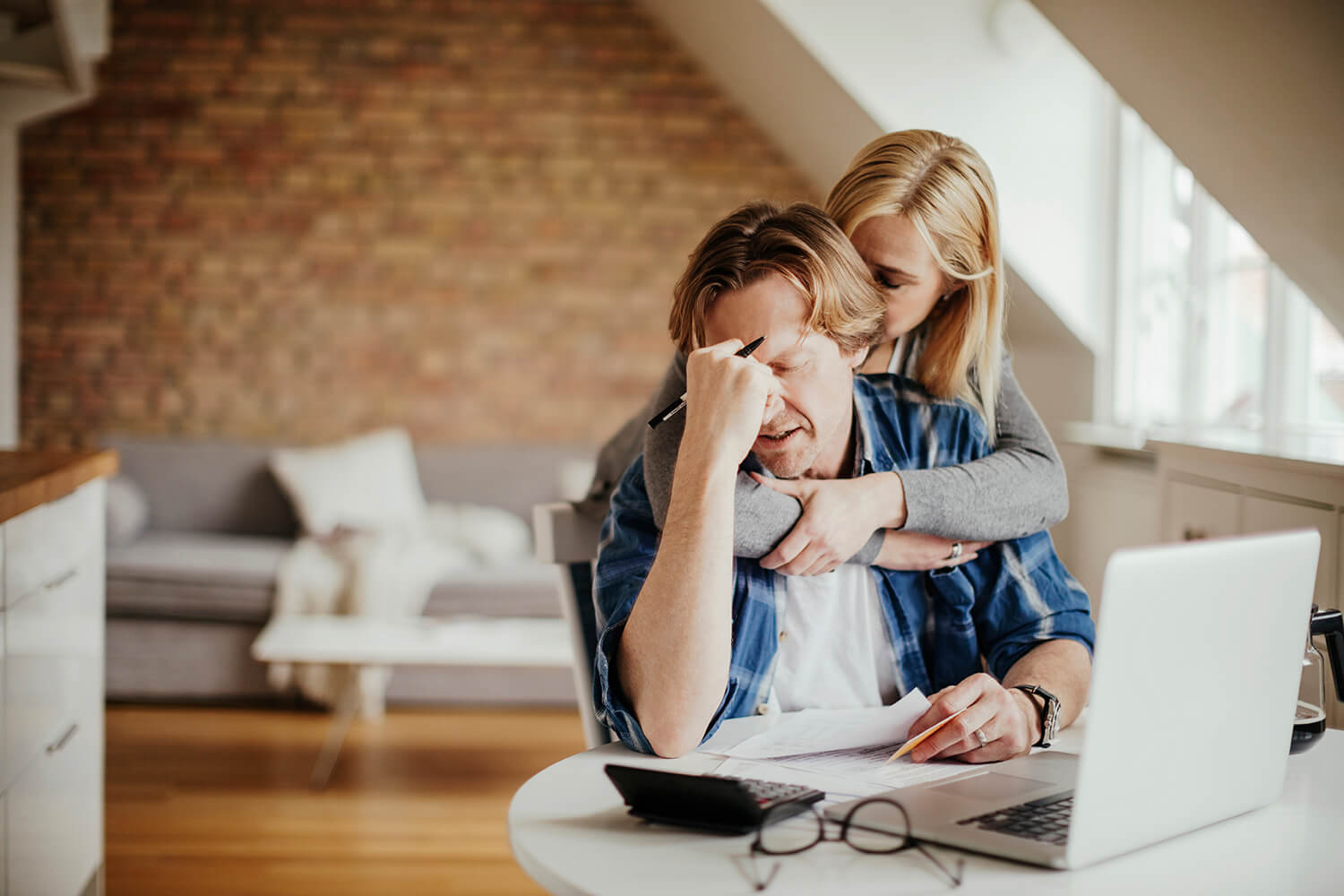 Frustrated, a man sitting at a table with a laptop and papers rubs his eyes while a woman hugs him. If you're denied Social Security Disability in Alabama, you can appeal the decision.