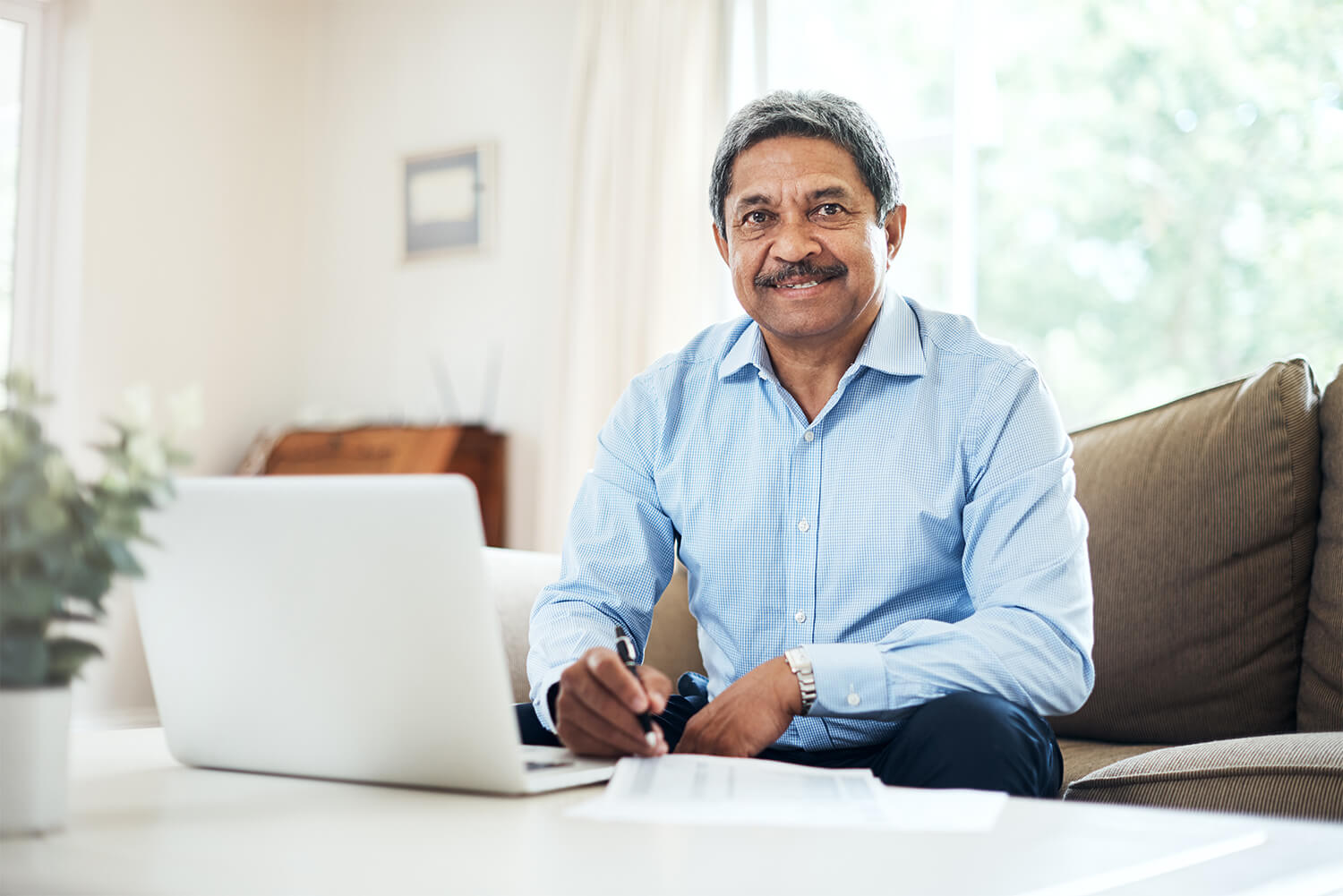 A man sits on a couch with a laptop on the coffee table, holding a pen and smiling. The Clarkson Firm helps people in Alabama get Social Security Disability Insurance (SSDI) benefits.