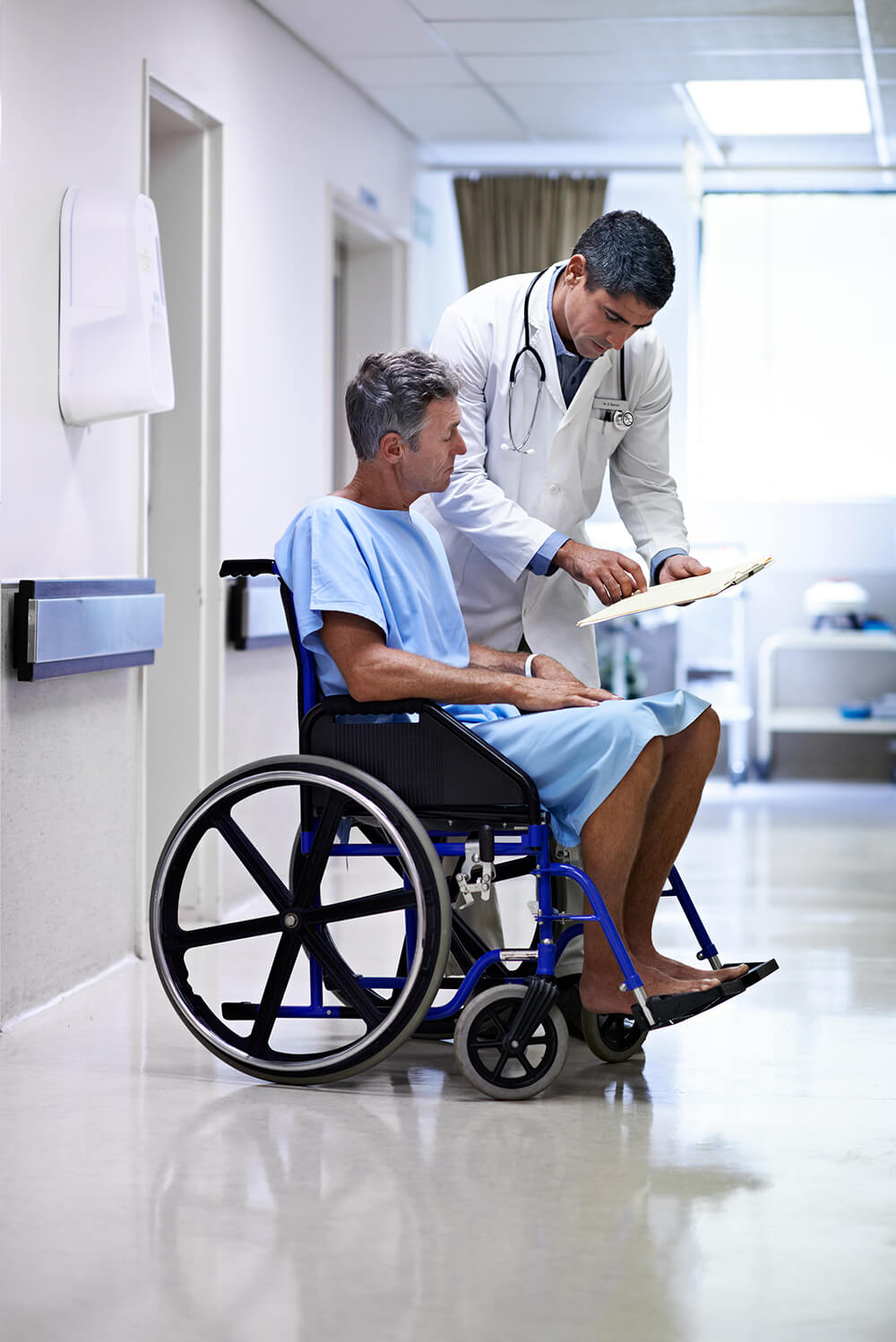 Wearing a hospital gown and sitting in a wheelchair in a hospital hallway, a man listens as a doctor stands over him showing him information on a clipboard. The Clarkson Firm helps people in Alabama with personal injury cases and civil litigation.