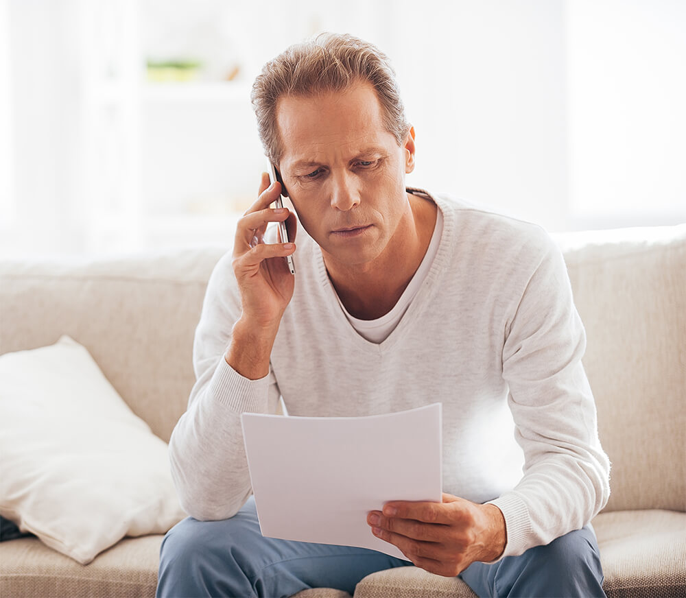 Looking concerned, a man sits on his couch, reading from a sheet of paper and talking on the phone. The Clarkson Firm disability attorneys can answer questions about getting Social Security Disability benefits.